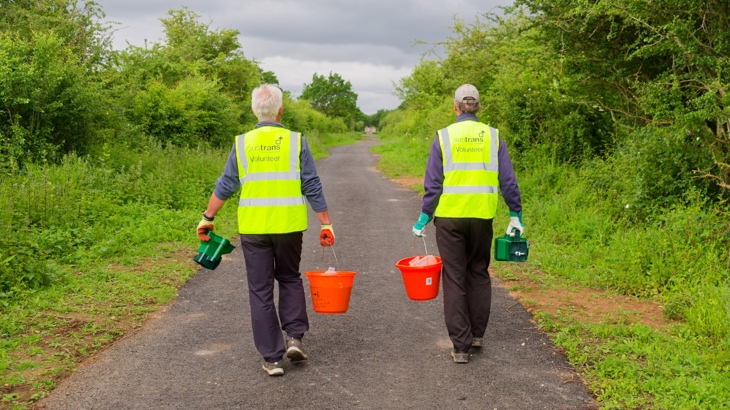Two Walk Wheel Cycle Trust volunteers wearing high visibility jackets walking along a path holding red buckets with wildflower seedings in.