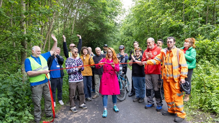 a crowd stands on a cycle route cheering on a woman in a red coat cutting a ribbon.