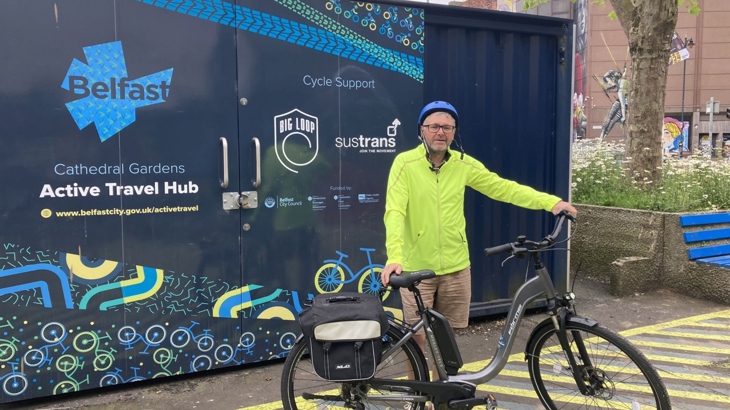 A man wearing a helmet and hi-vis jacket stands with an electric bicycle outside an active travel hub in Belfast city centre.
