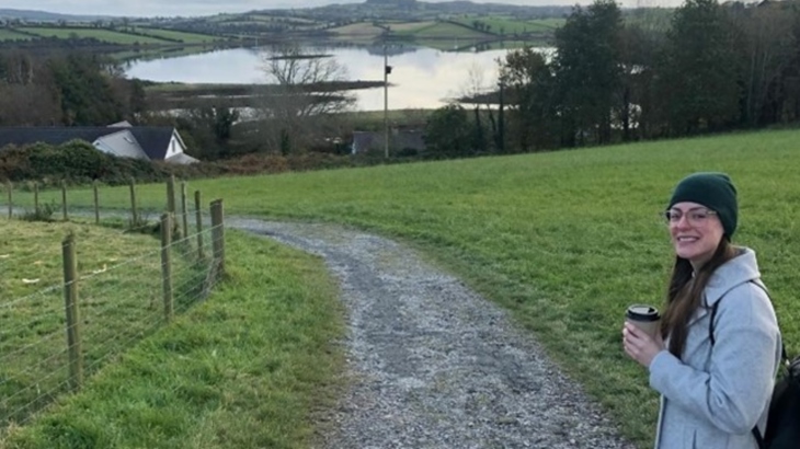 A woman wearing a hat and glasses turns to smile at the camera while walking along a country path with a lake in the background.