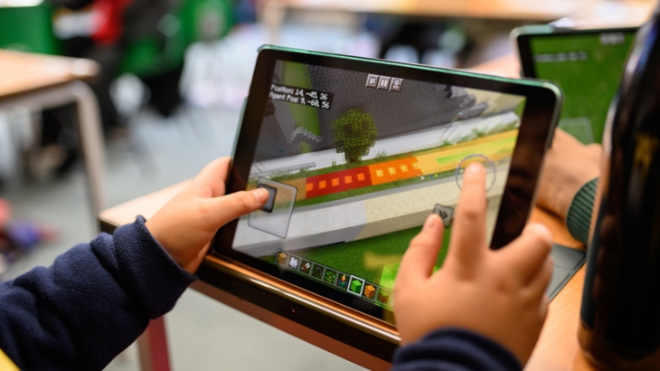 A child's hands hold an iPad showing a street design scene in the game Minecraft