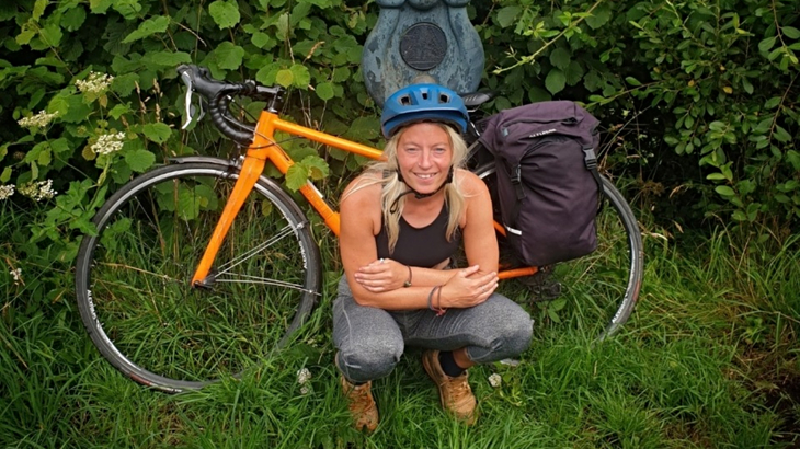 A woman with blonde hair wearing a helmet crouching down next to her orange bike and a Walk Wheel Cycle Trust's Millennium Milepost