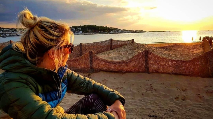A woman wearing sunglasses and a puffer jacket sat on a beach in Devon looking out at the setting sun on the sea