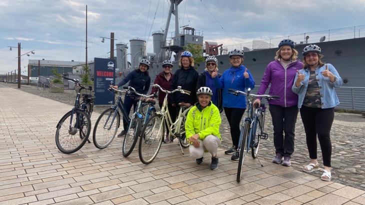 A group of women wearing helmets with bikes stand in front of a grey ship at a slipway in the Titanic Quarter of Belfast.