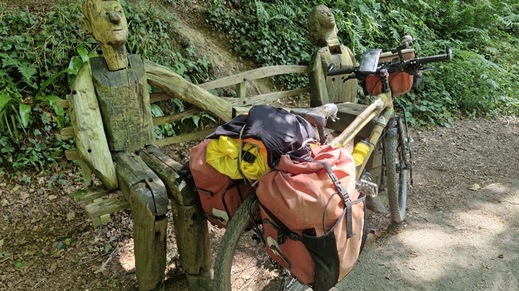 Two figures carved out of wood on a bench, with the bamboo bike standing in front of it.