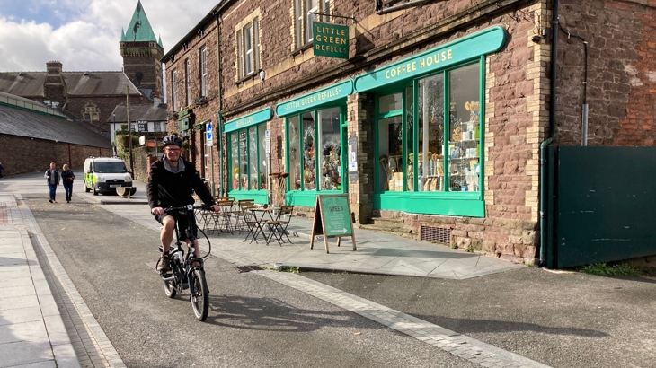 Abergavenny resident Daniel riding past the Little Green Refills shop in Abergavenny a black e-bike on a sunny day