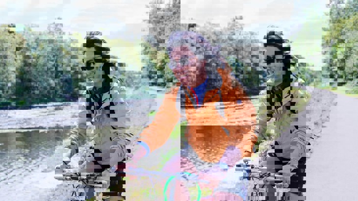 Cyclist in dark glasses and orange jacket on asphalt cycle path by river