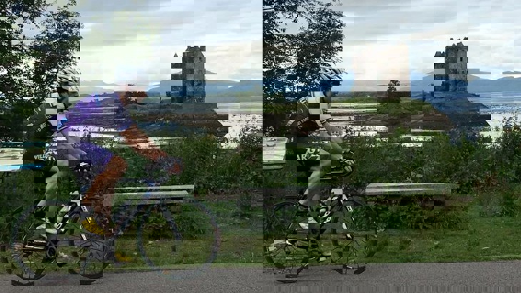 Man cycling on road bike on asphalt path in white helmet and blue and white lycra. To the side is loch with island and castle on it