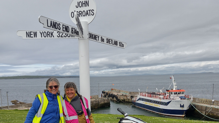 Two women stood smiling both wearing high-vis vets next to the John O'Groats sign in Scotland by the coast on a cloudy day