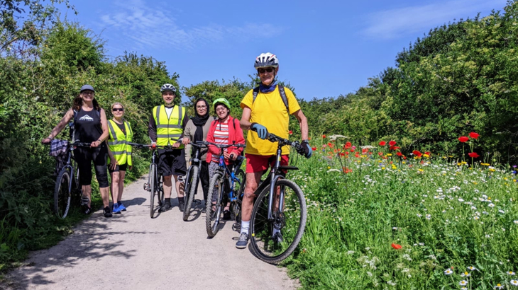 A group of volunteers on their bikes stand next to their flowering patch of wildflowers on a sunny day.