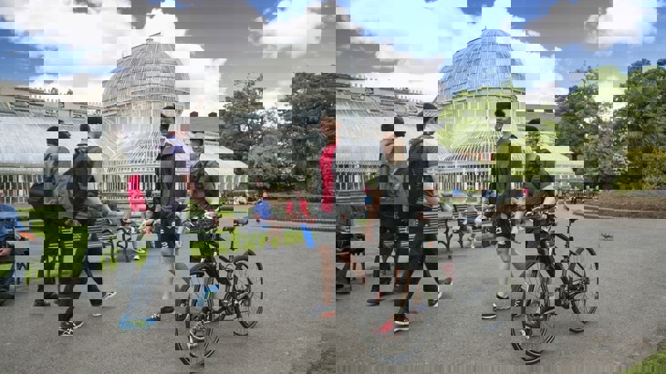 Two young men, one pushing a bike, are walking past The Palm House in Botanic Gardens, South Belfast on a sunny, blue sky and fluffy white cloud day. Other people are walking in the background and sat on benches enjoying the mild day.