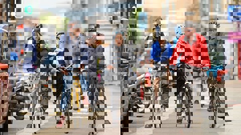 Three adults wearing helmets and riding bikes are waiting at a traffic light in a cycle lane in a city, there are queuing cars beside them and everyone is smiling.