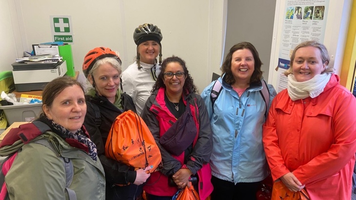 Six women, two wearing bicycle helmets and three holding orange bags, stand in a room, smiling at the camera.