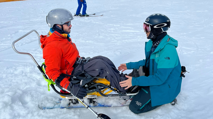 A man in a sit-ski wearing a helmet and a red jacket facing a ski instructor who is sat in front of him wearing a blue coat and a helmet