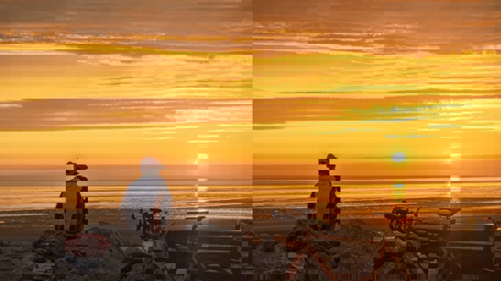 A cyclist sitting on some rocks looking out to sea as the sun sets, with poppy wreath and bike leaning up against a bench