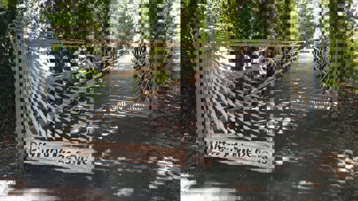 A pair of decorative gates are set at a staggered distance on a traffic-free path. The gates are a pale, galvanized metal. They have sweeping lines which end in a scroll. The words 'Dyffryn Maes Glas' are cut out of the bottom of each gate. The path beyond is long, straight and tree-lined, with a wooden fence running either side.