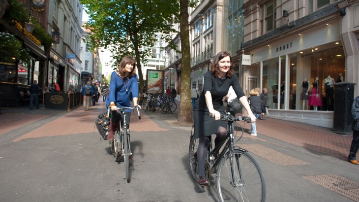 Two women riding bikes through a town centre