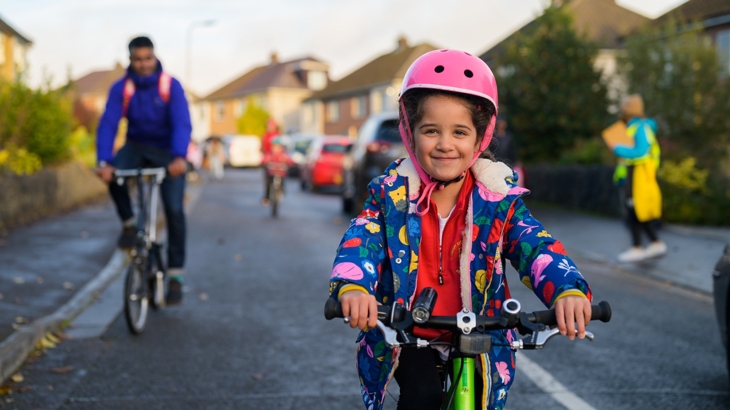 A small girl riding her bike in the foreground, with people cycling behind her in the background on a residential street.