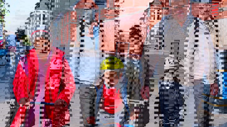 Family with dad walking, daughter scooting and son cycling to school together in the sunshine