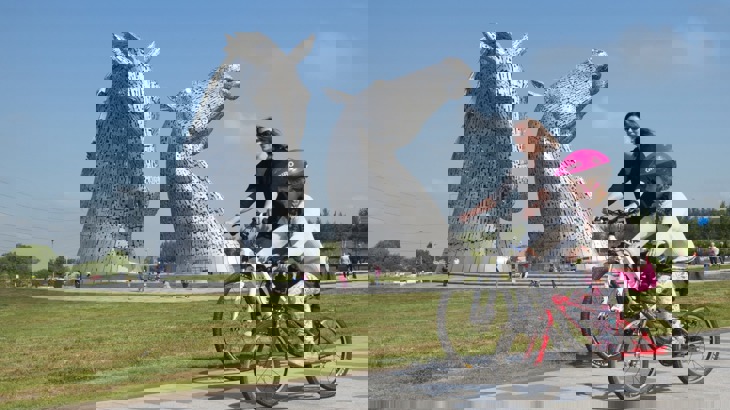 Young girl and woman riding cycles on path near giant horse head sculptures