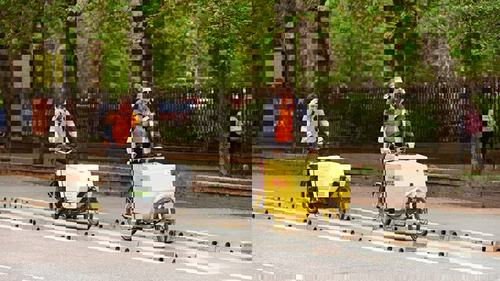 Two men riding cargo bikes along a segregated cycle lane in Manchester, past a tree-lined park.