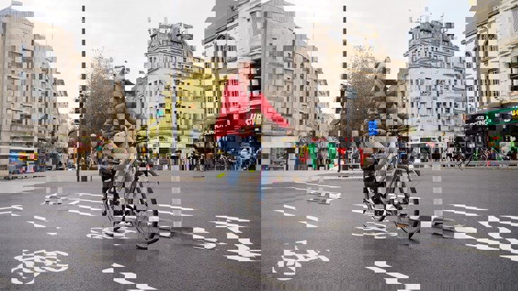 Woman wearing a red waterproof smiling as she cycles along a new temporary cycle lane in a city centre.