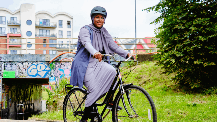 A woman wearing her Hijab under a cycle helmet is posing with a bike in the city. In the foreground is grass and in the distance street art on a bridge and modern flats.