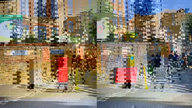Red sign next to a red postbox, showing that the street outside the school gate is closed to vehicle traffic, as children play and laugh in the street. Healthy Streets Officers London