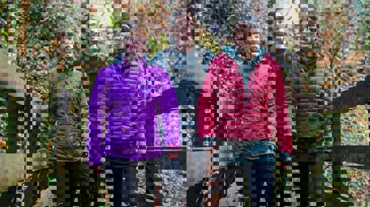 Three friends wearing brightly coloured waterproof jackets walking along a bridge chatting, surrounded by orange autumnal leaves and trees.