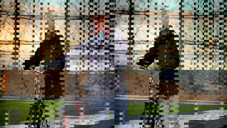 Lady wrapped up in a black fleece coat, scarf and gloves stands smiling with her bicycle next to her.