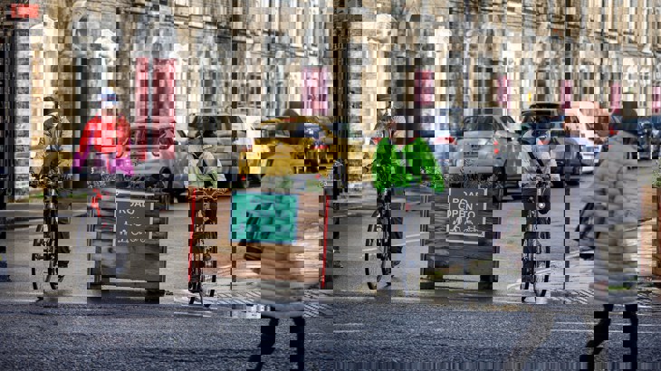 Two friends with bicycles stop at the end of a road next to a sign which reads "Road open to" followed by symbols for a parent and child, push scooter, wheelchair and bicycle.