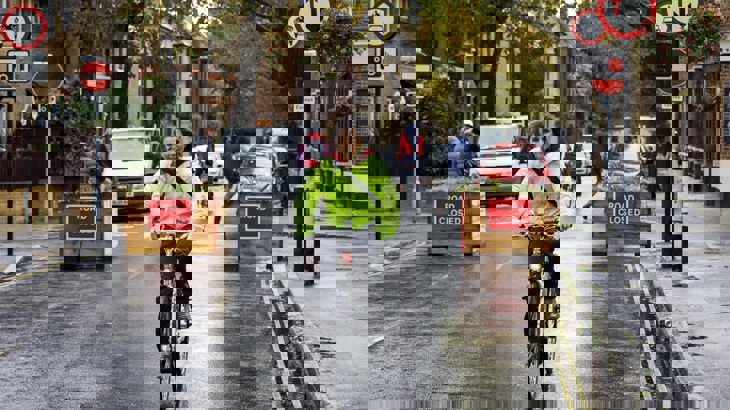 A lady rides her bike on a road with no moving cars. There are two planters behind sayin 'road closed'. It is a wet, autumn day.