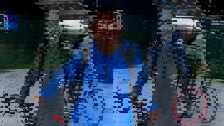 Girl wearing a hoodie stands with her bicycle smiling at the camera whilst a man cycles past in the background.