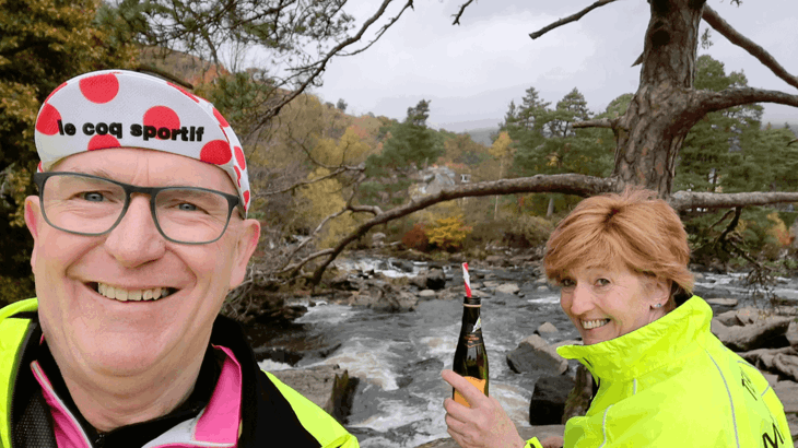 Colin takes a selfie with Diane in the background, holding a bottle of prosecco. Both are in neon cycling gear overlooking a river. Colin wears an excellent spotty hat.