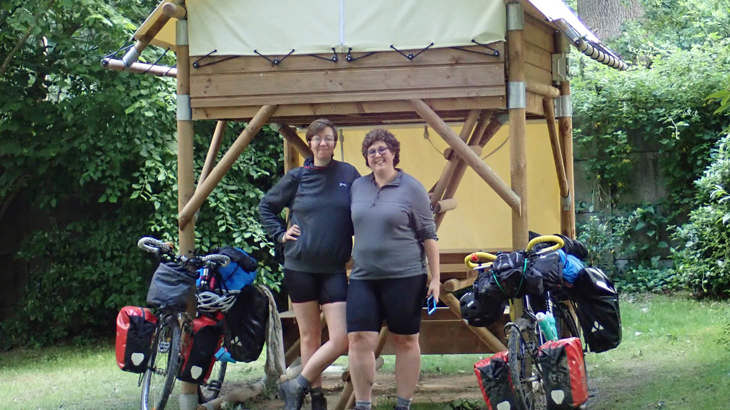 Abi and Lilith standing together smiling with their cycle touring kit and bikes, in front of a tree house.