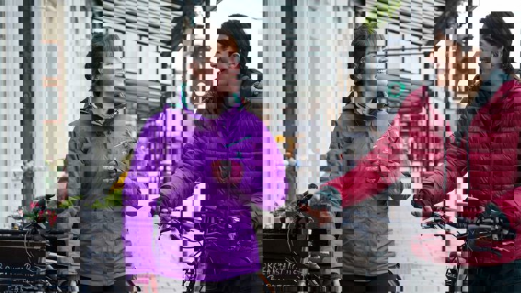 Two women wearing brightly coloured raincoats, chatting and walking with their bikes through a high street