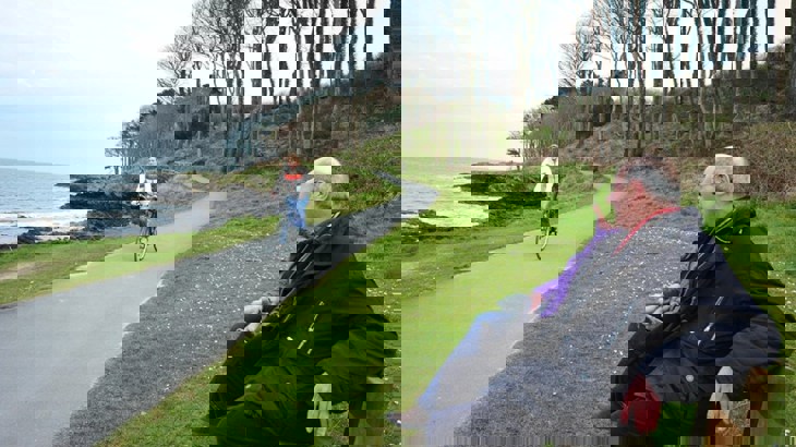 A cyclist on the North Down Coastal Path watched by two people sitting on a bench 