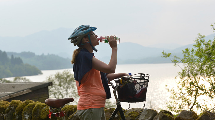 Lady standing with bike in the sunshine. She is wearing helmet and drinking from a reusable bottle.