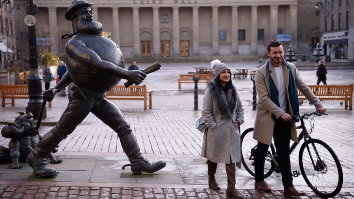 A woman wearing a hat and coat walks beside a man wearing a blazer and pushing a bike in Dundee. A statue is to the left of them.