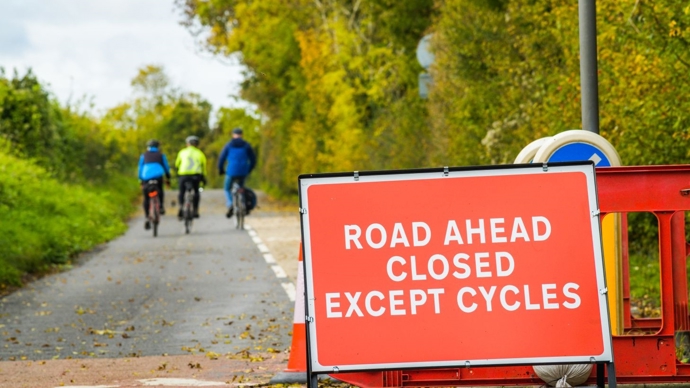 Road sign in forefront reads 'Road ahead closed except cycles', three adults cycle along the road in the background, road lined by trees