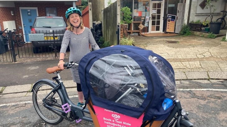 Pregnant woman on a cargo bike in a side street. The bike is loaded with equipment and has a rain cover.