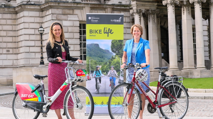 Belfast Lord Mayor Kate Nicholl standing with a Belfast Bike and Walk Wheel Cycle Trust Caroline Bloomfield standing with her bike in front of Belfast City Hall with a Bike life pop up display stand showing two females cycling in North Belfast
