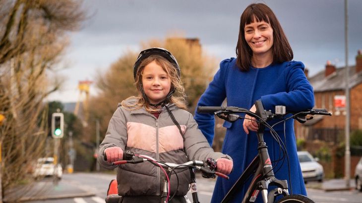 Little girl wearing helmet holds her bike next to female politician with her bike, beside a road.