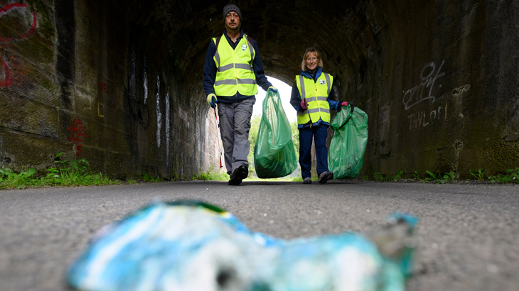 Two volunteers with litterpickers approach a discarded tin can along a traffic-free National Cycle Network path in Ayrshire.