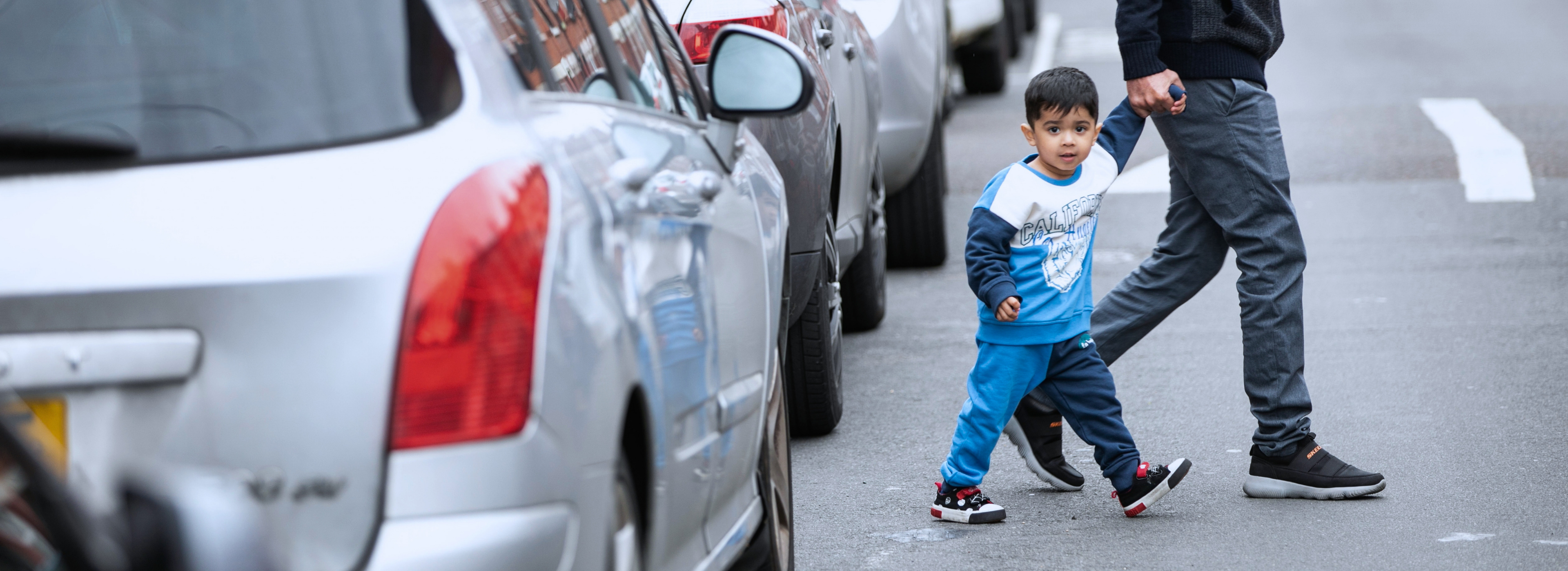Little boy crosses a city road, lined with parked cars, holding his dad's hand and looking earnestly at the camera