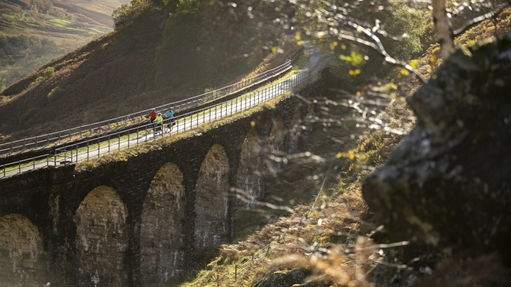 An aerial shot of three adults cycling over Glen Ogle Viaduct on the National Cycle Network Route 7; showing heather and autumn colours in the background.