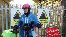 A person standing in front of a closed off route with their electric bike. Behind them is a sign which reads "footway closed".