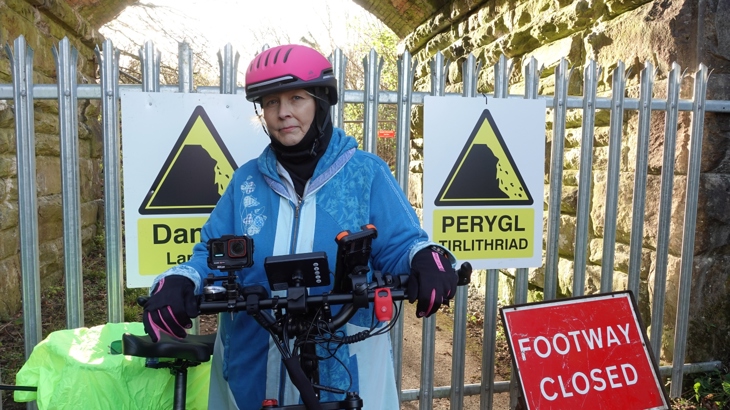 A person standing in front of a closed off route with their electric bike. Behind them is a sign which reads "footway closed".