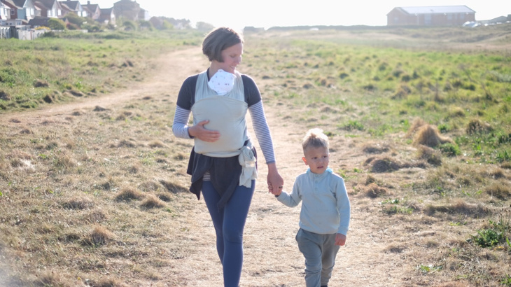 A woman with a newborn baby in a sling around her body walking on a sandy path with grass either side holding the hand of her young son on a bright day