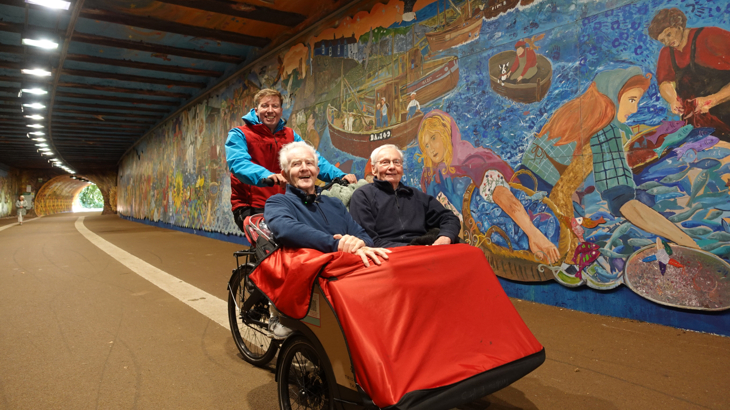 A trishaw with two passengers smiling and Mark, the trishaw pilot on the bike, in front of the Alloway Tunnel art mural.
